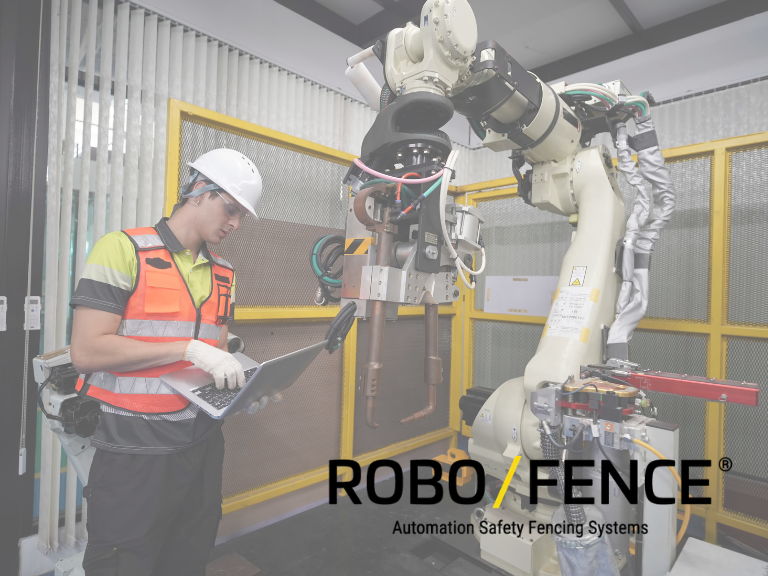 A person is holding and looking down at a laptop while standing next to a cobot with fencing behind it